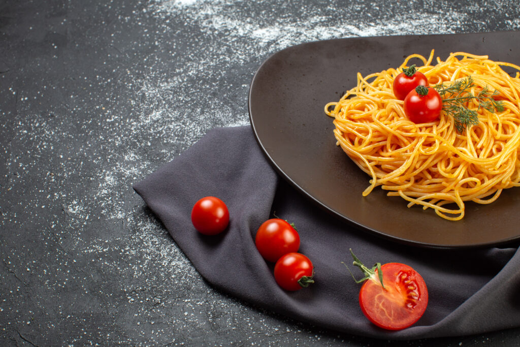 vorderansicht-von-koestlichen-spagetti-auf-einem-schwarzen-teller-auf-einem-dunklen-farbtuch-rohe-nudeln-frische-tomaten-auf-der-linken-seite-auf-einem-dunklen-farbtisch